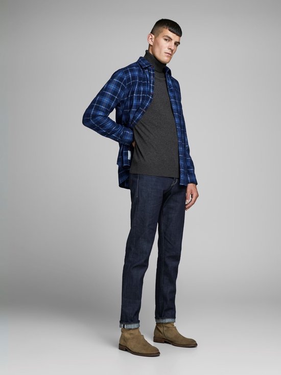 Man in donkergrijs coltrui, met open blauw-zwart geruite blouse en spijkerbroek.