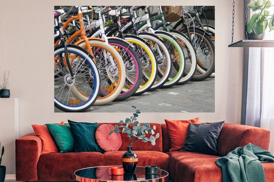 Une rangée de vélos colorés avec des jantes orange, bleues, blanches, roses et jaunes, au-dessus d'un canapé rouge dans un salon.