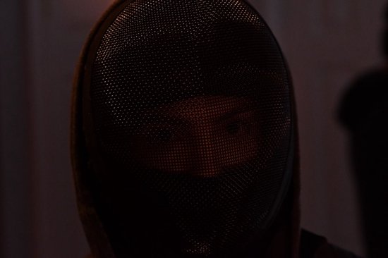 Close-up van een zwaardvechtersmasker dat een oog zichtbaar laat, donkere sfeer.