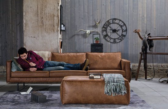 Een man ligt op een cognac leren bank met een plaid en een voetenbankje.
