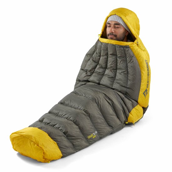Man in grijze muts zit rechtop in een donkergrijze en gele slaapzak.