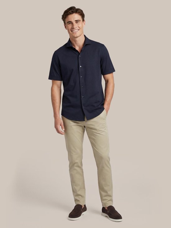 Man in donkerblauw overhemd met korte mouwen en beige chino broek, staand met hand in broekzak.