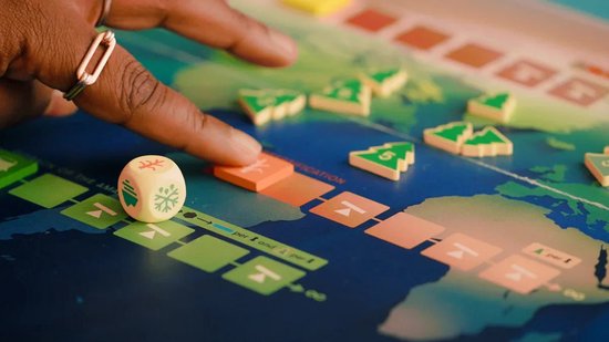 Een hand met ring speelt het bordspel Daybreak. Een dobbelsteen met een sneeuwvlok ligt op het spelbord. Het spelbord toont een wereldkaart.