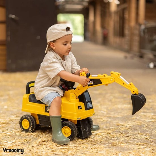 Gele loopauto graafmachine met een jongen met een pet en groene laarzen, zittend op stro.