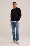 Man staand, dragend een zwarte sweater en een blauwe jeans met lichte wassing.