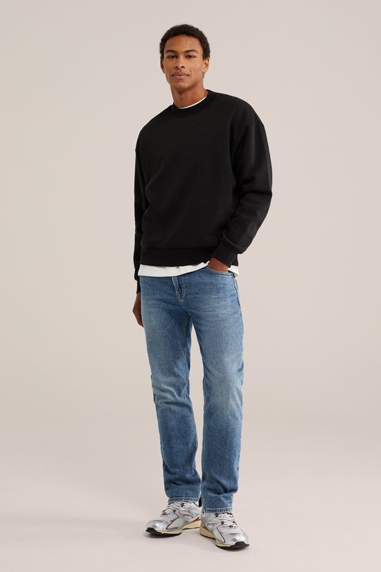 Man staand, dragend een zwarte sweater en een blauwe jeans met lichte wassing.