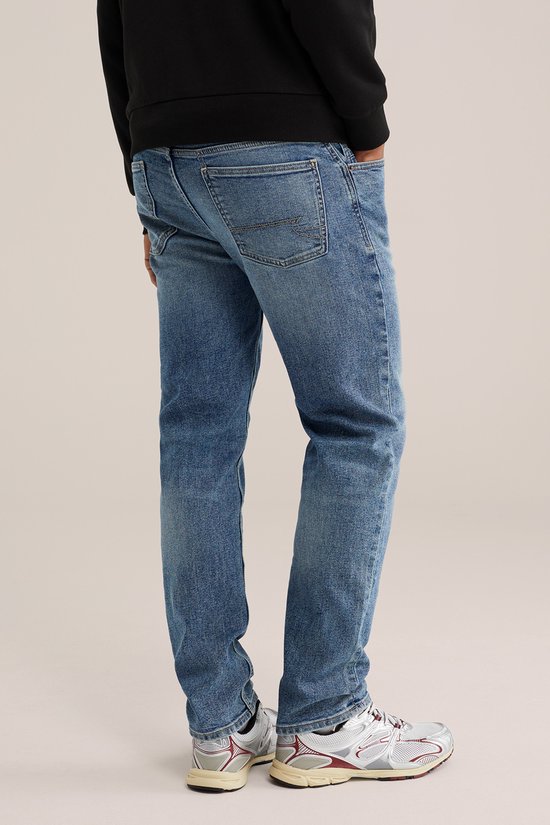 Man die een zwarte sweater en een blauwe jeans met lichte wassing draagt, gezien vanaf de zijkant.