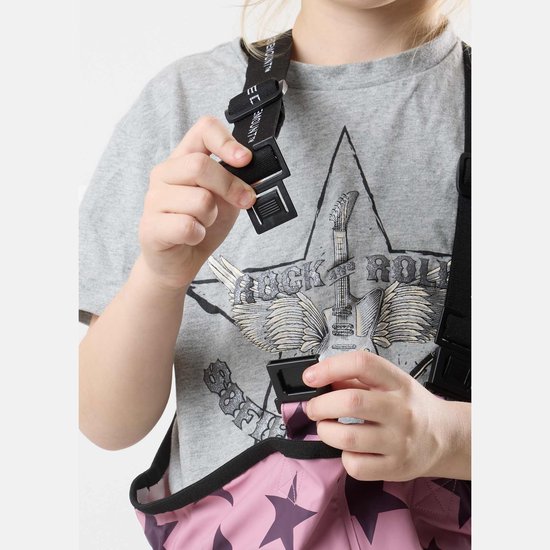 Meisje met bril en een grijze t-shirt met rock 'n roll print, dat de sluiting van de roze, sterrenbedrukte regenbroek vastklikt.