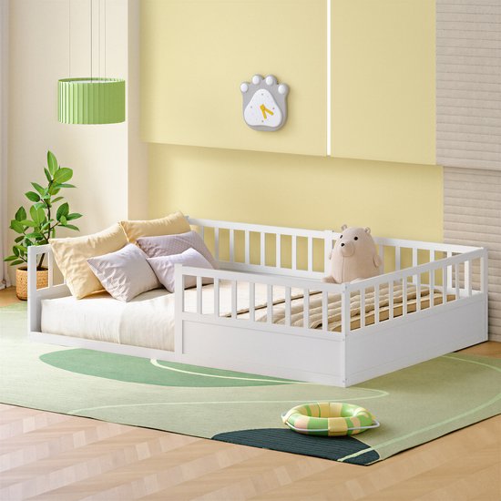 Wit kinderbed met ombouw en kussens, een knuffelbeer en een speelgoedring op een groene mat.