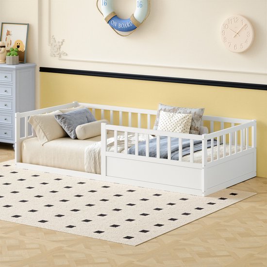 Wit houten peuterbed met uitvalbeveiliging, opgemaakt met kussens en dekens, in een kinderkamer met gele muur.