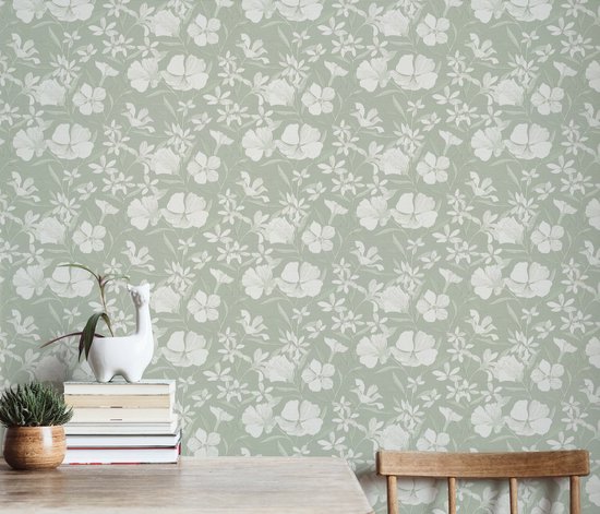 Lichtgroen behang met een delicaat wit bloemen- en bladerenpatroon, getoond op een muur met een houten tafel.