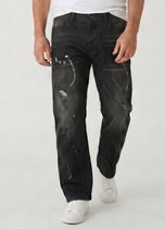 Een man draagt een zwarte, baggy spijkerbroek met witte verfspatten, gezien vanaf de voorkant.