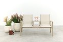 Beige aluminium tuinbank met stoffen zittingen, middenplateau met twee roze glazen, naast grote planten.