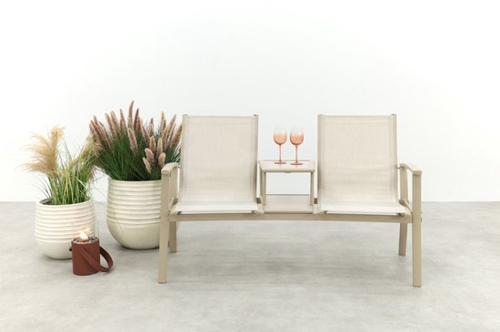 Beige aluminium tuinbank met stoffen zittingen, middenplateau met twee roze glazen, naast grote planten.