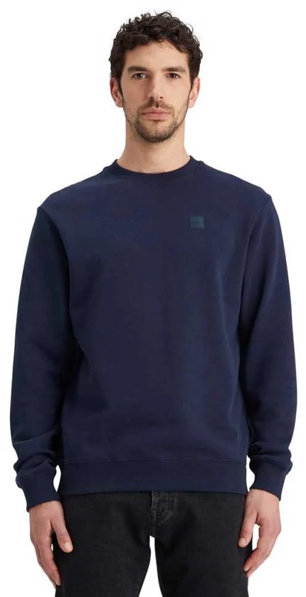 Man met donkerblauwe sweater met ronde hals en een klein ruitvormig logo op de borst.