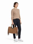Vrouw met bruine suède tas met houten handvatten en bruine franjes. Ze draagt een beige trui en zwarte broek.