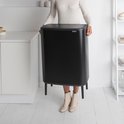Une femme en pull blanc à col roulé et bottines beiges soulève une grande poubelle Brabantia noire sur quatre pieds.