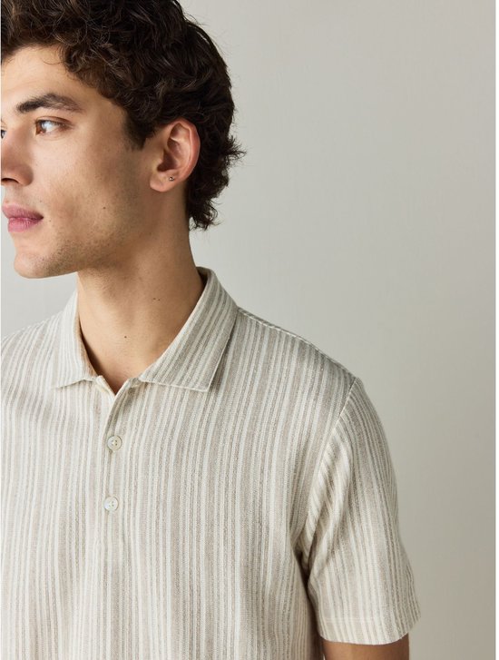 Zijaanzicht van een man in een beige polo met fijne verticale strepen en een klein zilveren oorbellen.