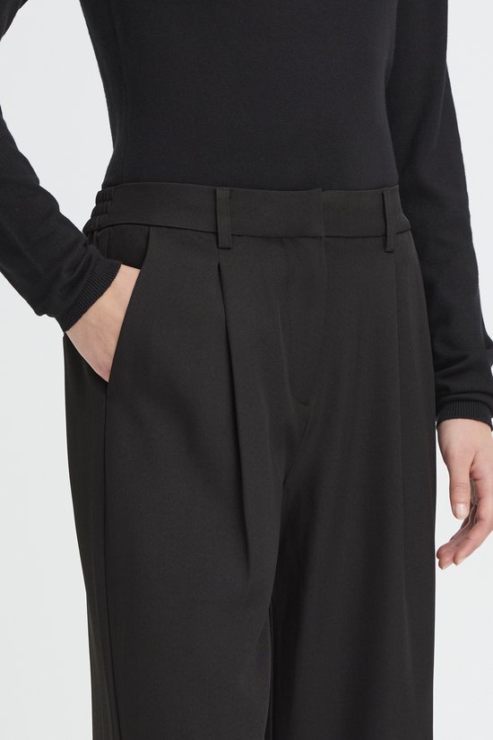 Detail van de voorkant van een zwarte wijde broek met plooien en steekzakken.