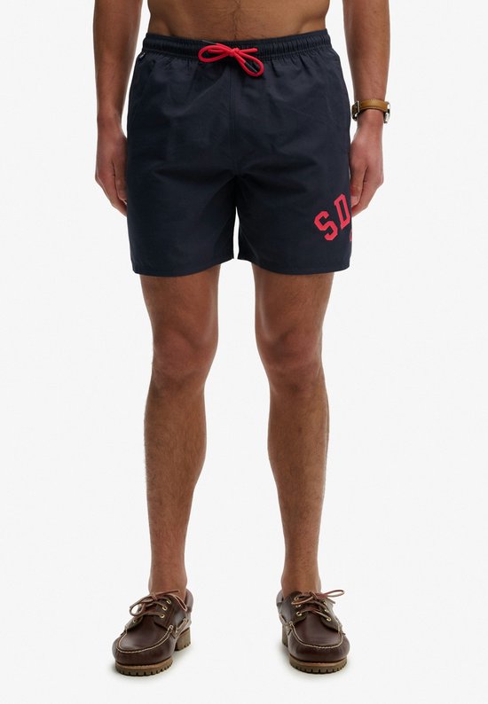 Un homme portant un maillot de bain bleu marine avec l'inscription rouge 'SD' sur la jambe gauche.