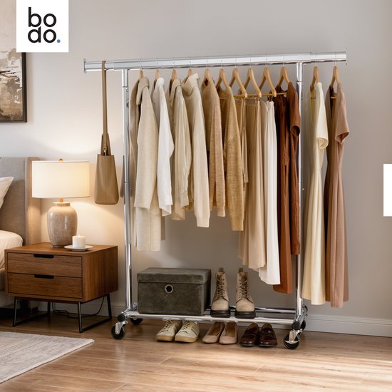 Zilveren kledingrek met kleding in neutrale tinten. Op de nachtkastje staat een lamp, naast het rek een plant.