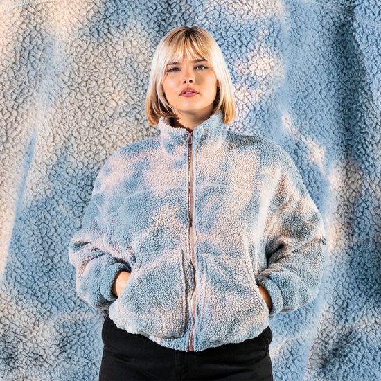 Model met blonde bob draagt een blauw met witte teddy jas met rits. Achtergrond is een zachtblauwe, fluffy stof.