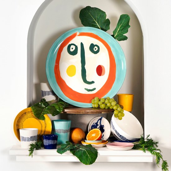 Een serveerschaal met een kindertekening van een gezicht, met een blauwe rand, oranje haar en gele en rode wangen, in een nis.