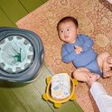 Baby in blauw rompertje ligt naast een luiertoren in een gele schildpadmand, met een grijze luieremmer met groene zakken ernaast.