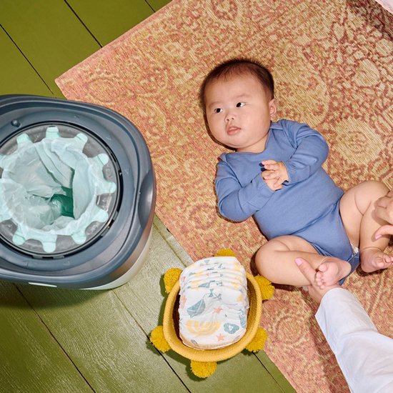 Baby in blauw rompertje ligt naast een luiertoren in een gele schildpadmand, met een grijze luieremmer met groene zakken ernaast.