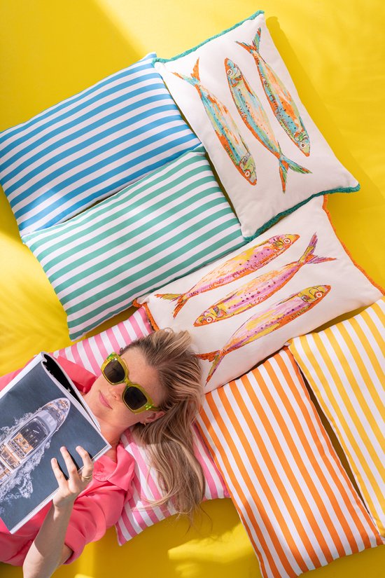 Een vrouw ligt op een gele achtergrond, omringd door gestreepte kussens in blauw, groen, oranje en geel. Ze houdt een tijdschrift vast en ligt tussen kussens met sardines erop.