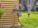 Gardena waterverdeler met kraan en digitale timer. Twee slangen verbonden, één leidt naar een sproeier in de tuin.