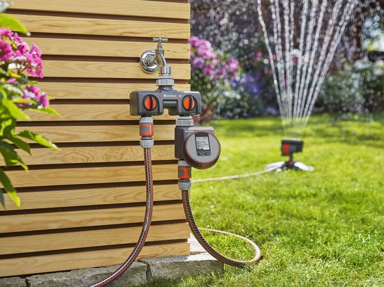 Gardena waterverdeler met kraan en digitale timer. Twee slangen verbonden, één leidt naar een sproeier in de tuin.