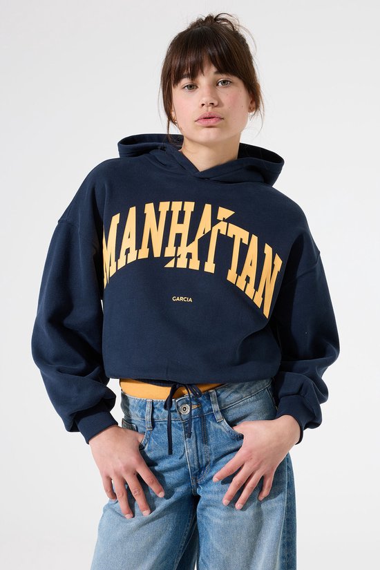 Mannequin portant un sweat à capuche court bleu marine avec inscription 'MANHATTAN' jaune et jean large.