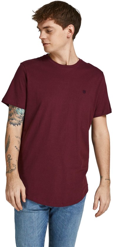 Man met tatoeages op zijn arm draagt een bordeauxrode t-shirt met korte mouwen en ronde hals, met een klein logo op de borst, staand.