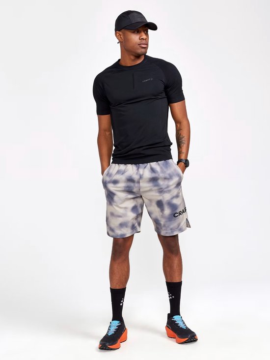Man in zwart Craft shirt met korte mouwen en grijze shorts met tie-dye patroon. Draagt zwarte pet en sportschoenen.