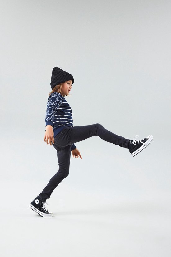 Jongen met zwarte beanie en gestreepte trui in een grijze slim fit spijkerbroek, beweegt met één been omhoog.