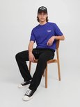 Model zittend op een houten stoel, draagt een zwarte pet met 'NEW YORK', blauw T-shirt en zwarte joggingbroek.