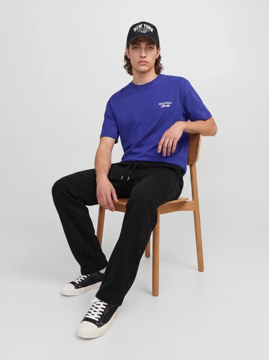 Model zittend op een houten stoel, draagt een zwarte pet met 'NEW YORK', blauw T-shirt en zwarte joggingbroek.