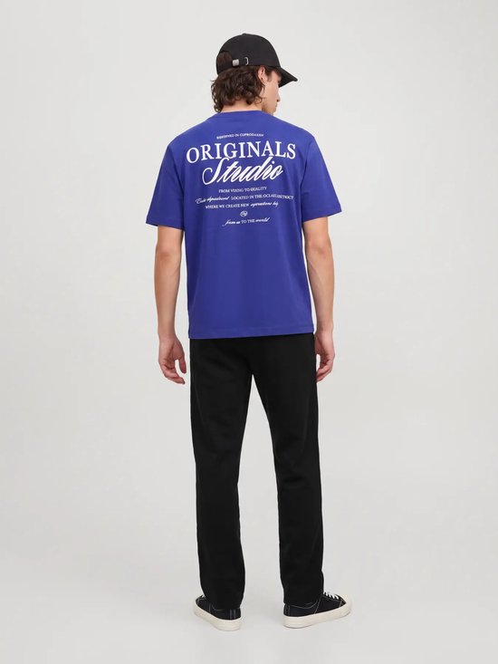 Achteraanzicht van model in blauw T-shirt met grote witte 'ORIGINALS Studio' tekst op de rug en zwarte broek.