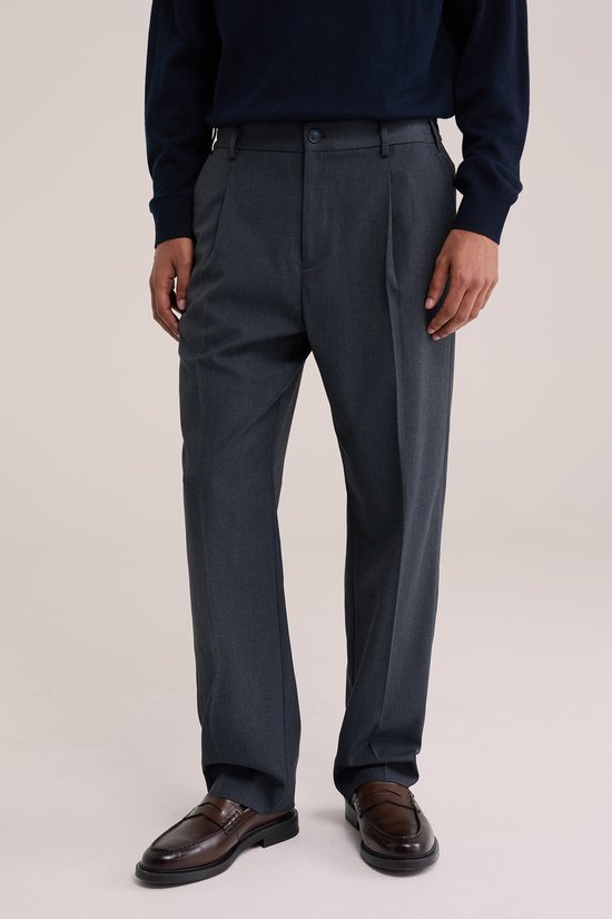 Man in donkerblauwe trui en donkerblauwe pantalon met bandplooi, met de handen in de zakken.