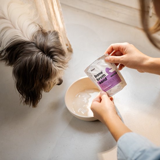 Een hond kijkt naar een pakje bottenbouillon van eend dat wordt geopend boven een voerbak.