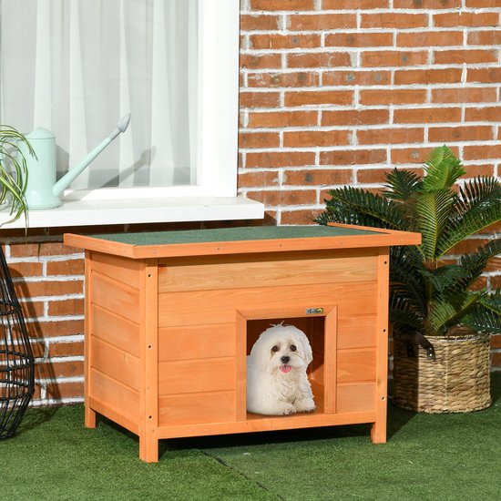 Wit hondje kijkt vanuit de ingang van een bruin houten hondenhok met groen dak.