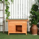 Bruin houten hondenhok met groen dak in een tuin, naast een ladder en planten.