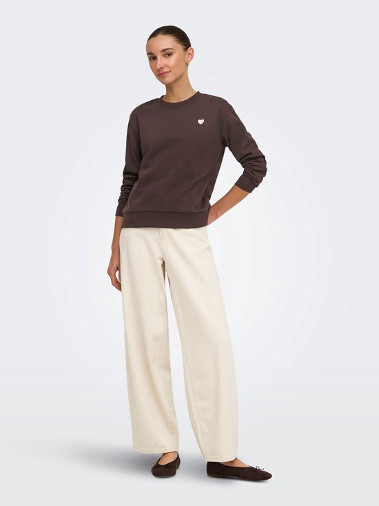 Vrouwenmodel met een donkerbruine trui met lange mouwen en een wit hartje op de borst. Gepaard met een crème broek.