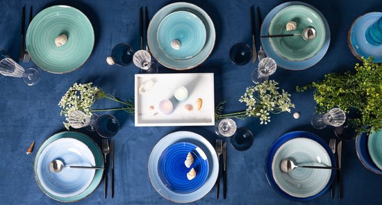 Een bovenaanzicht van een blauw gedekte tafel met keramische kommen in verschillende blauwtinten en schelpen.