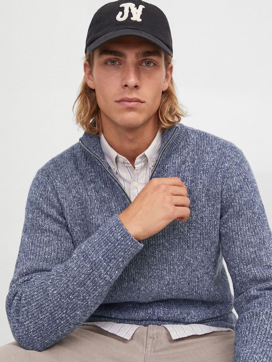 Model met lang blond haar draagt een blauw melange gebreid poloshirt met halve rits, gestreepte blouse en zwarte pet.