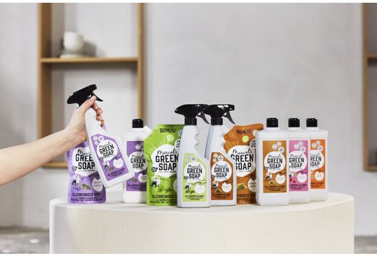 Hand houdt een Marcel's Green Soap sprayflacon vast voor een assortiment navulverpakkingen en flessen op een voetstuk.