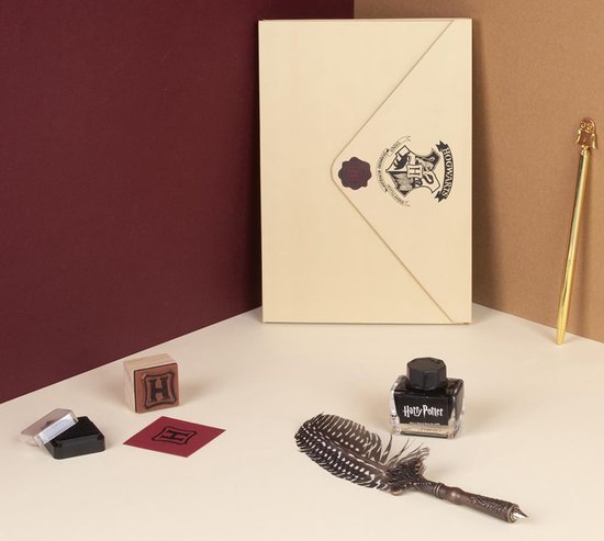 Harry Potter schrijfset: beige envelop met Hogwarts-zegel, veerpen, gouden pen, inktpot en H-stempel op een tafel.