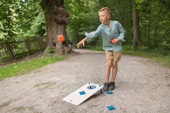 Jongen gooit oranje beanbag naar houten cornhole board met blauwe beanbags, in bosrijke omgeving.
