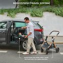 Man plaatst een beige Kinderkraft Mink Pro autostoel in een auto, naast de beige Esme Pro kinderwagen.
